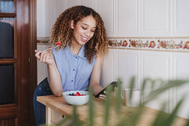 Schöne Frau, die ein Mobiltelefon benutzt, während sie in der heimischen Küche einen Obstsalat isst - EBBF03827