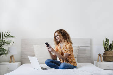 Lächelnde Frau mit Smartphone auf dem Bett zu Hause - EBBF03832