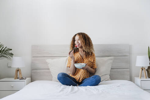 Junge Frau isst Obstsalat, während sie mit gekreuzten Beinen auf dem Bett sitzt - EBBF03840