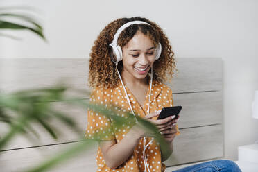 Junge Frau, die ein Mobiltelefon benutzt, während sie auf dem Bett zu Hause Musik hört - EBBF03848