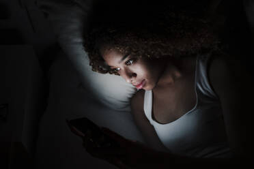 Woman using smart phone while lying on bed in dark room at home - EBBF03850