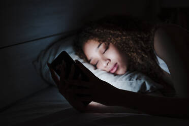 Frau hält Handy, während sie auf dem Bett in einem dunklen Raum zu Hause schläft - EBBF03851