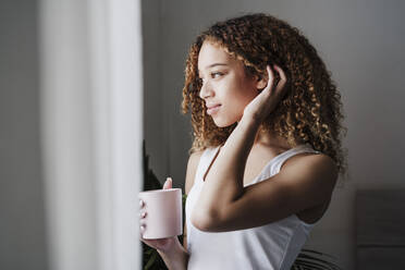 Schöne Frau mit Hand in den Haaren schaut weg, während sie im Schlafzimmer Kaffee trinkt - EBBF03852