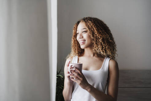 Lächelnde junge Frau, die durch ein Fenster schaut, während sie zu Hause eine Kaffeetasse in der Hand hält - EBBF03853