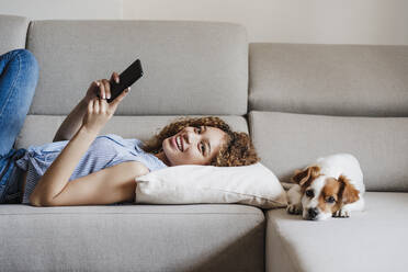 Lächelnde Frau mit Handy, die neben einem Jack Russell Terrier auf dem Sofa zu Hause liegt - EBBF03858