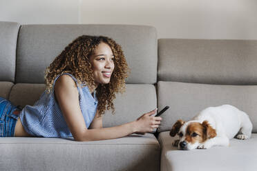 Lächelnde junge Frau mit Smartphone auf dem Sofa liegend neben Jack Russell Terrier zu Hause - EBBF03861