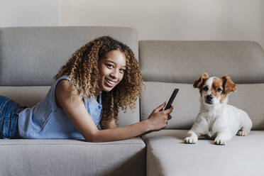 Curly haired woman with smart phone lying on sofa by dog in living room - EBBF03862