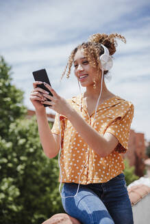 Frau, die ein Selfie mit ihrem Smartphone macht, während sie auf dem Balkon Musik hört - EBBF03874