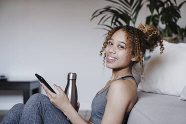Beautiful smiling woman with mobile phone and bottle leaning on sofa at home - EBBF03881