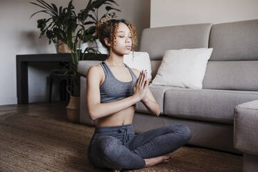 Junge Frau mit verschränkten Händen beim Meditieren auf einem Teppich zu Hause sitzend - EBBF03896
