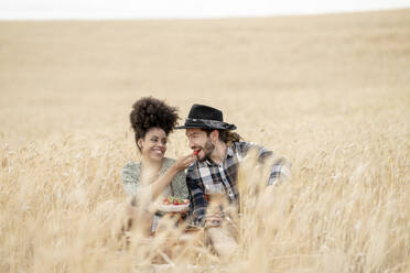 Cheerful girlfriend feeding strawberry to boyfriend while sitting on field - JCCMF02775