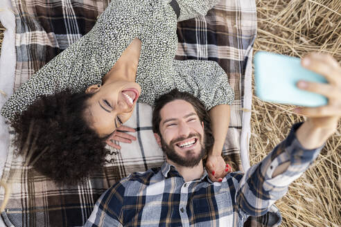 Fröhlicher Freund nimmt Selfie durch Smartphone mit Frau, während auf Decke liegend - JCCMF02785