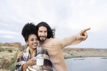 Boyfriend pointing while standing by girlfriend wrapped in blanket - JCCMF02820