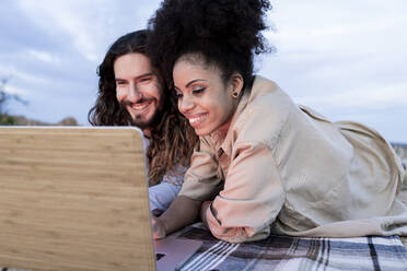Couple smiling while watching laptop during sunset - JCCMF02834