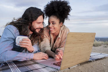 Mid adult couple smiling while lying by laptop on blanket during sunset - JCCMF02835