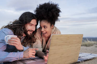 Boyfriend talking to girlfriend while lying by laptop during sunset - JCCMF02836