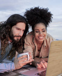 Smiling girlfriend and boyfriend watching laptop while lying on blanket - JCCMF02837