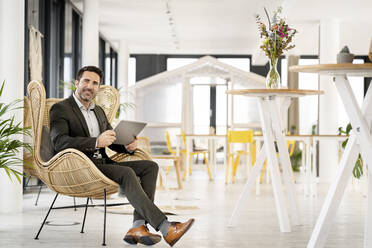Lächelnder männlicher Unternehmer mit digitalem Tablet, der auf einem Stuhl in einer Büro-Cafeteria sitzt - PESF02837