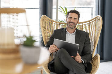 Lächelnder männlicher Fachmann, der ein digitales Tablet hält, während er auf einem Stuhl im Büro sitzt - PESF02840