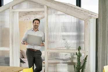 Smiling businessman with laptop leaning on doorway of cabin at office cafeteria - PESF02845