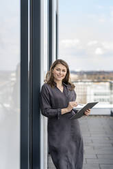 Lächelnde weibliche Fachkraft mit Grafiktablett am Fenster lehnend auf dem Balkon eines Büros - PESF02877