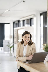 Smiling businesswoman leaning on table in front of laptop at cafeteria - PESF02914