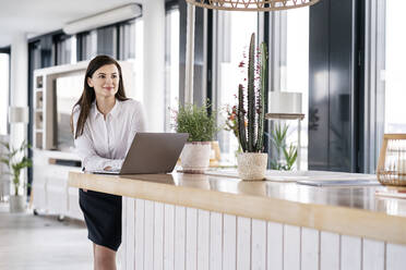 Geschäftsfrau mit Laptop, die sich in einer Büro-Cafeteria an den Tresen lehnt - PESF02916