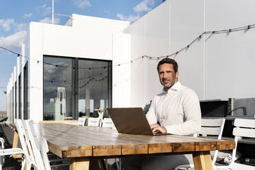 Businessman with laptop sitting at table on office terrace - PESF02929