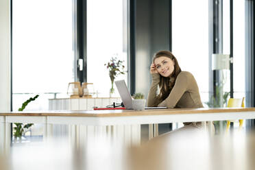 Lächelnde Geschäftsfrau mit dem Kopf in der Hand, die am Tisch im Büro sitzt und nachdenkt - PESF02936