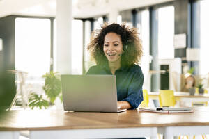 Smiling female professional talking through headset while working on laptop at office cafeteria - PESF02939