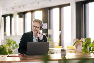 Male customer service representative with laptop looking away while talking through headset at cafeteria - PESF02948