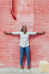 Cheerful woman with arms outstretched leaning on red brick wall - JRVF00944