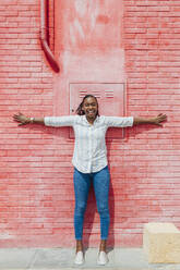Fröhliche Frau mit ausgestreckten Armen, die sich an eine rote Backsteinmauer lehnt - JRVF00944