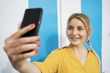 Smiling blond woman taking selfie through mobile phone by wall - RCPF01151