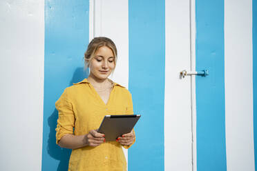 Blond woman using digital tablet in front of striped blue door - RCPF01154