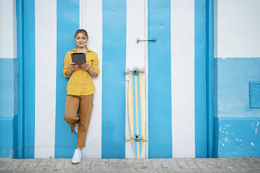 Smiling woman using digital tablet by longboard while leaning on striped door - RCPF01157
