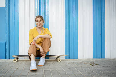 Beautiful woman sitting on longboard at footpath in front of striped wall - RCPF01160