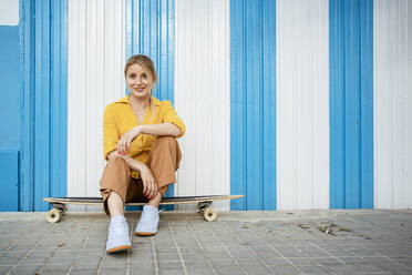 Schöne Frau auf Longboard sitzend auf Gehweg vor gestreifter Wand - RCPF01160