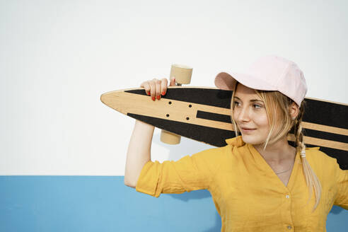 Smiling woman looking away while carrying longboard on shoulders in front of white wall - RCPF01162