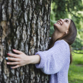 Lächelnde Frau umarmt Baum in öffentlichem Park - MCVF00881