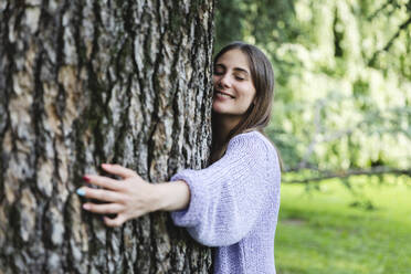 Smiling woman with eyes closed hugging tree in public park - MCVF00882