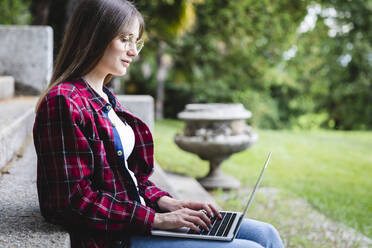 Junge Frau mit Brille benutzt Laptop in einem öffentlichen Park - MCVF00885