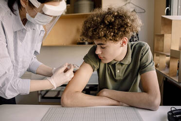 Boy with curly brown hair looking at nurse injecting medicine at home - MASF23817