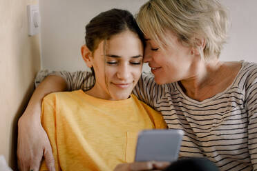 Happy mother embracing daughter using mobile phone in bedroom - MASF23836