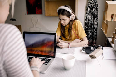 Mädchen mit Kopfhörern E-Learning über ein digitales Tablet, während sie mit einer Geschäftsfrau zusammensitzt, die an einem Laptop am Schreibtisch arbeitet - MASF23857