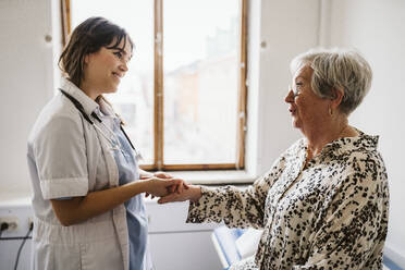 Smiling female healthcare worker looking at senior patient talking while holding hands - MASF23904
