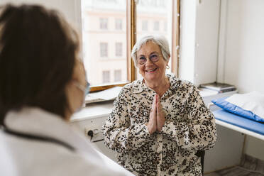 Lächelnder älterer weiblicher Patient mit verschränkten Händen, der den Arzt in einer medizinischen Klinik anschaut - MASF23907