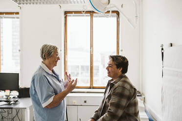 Ältere Ärztin gestikuliert während eines Gesprächs mit einem jungen männlichen Patienten in einer medizinischen Klinik - MASF23931