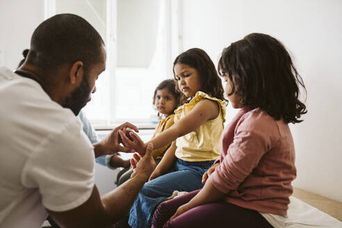 Male healthcare worker examining girl's hand sitting by sisters at medical clinic - MASF24024