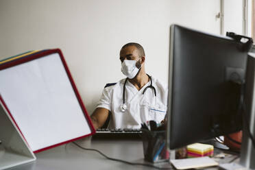 Male doctor wearing protective face mask reading medical files while sitting at desk - MASF24039